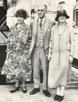 Three white people stand on a ship's deck for a photo: an older woman in a floral dress and dark hat, an older balding man in a suit, and a young woman in a cloche hat and dress with dark buttons down the front