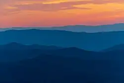 Five mountain ridges in shades of dark blue below an orange and yellow sunset.