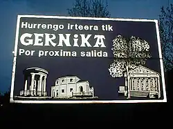 A flash photograph of a dark road sign with a white border. There is a line "Hurrengo irteera tik", another with "GERNIKA" in a bold font with heavy serifs and another with "Por proxima salida". Along the bottom and right borders there are depictions in black and white of a round columned kiosk, a church and a Classicist kiosk behind a tree.
