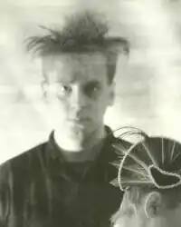 Man with dark hair wearing dark clothing against a plain background