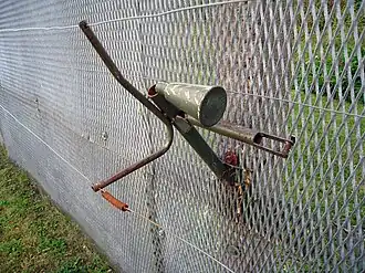 Horn-shaped device mounted on the side of a metal fence, with trigger wires attached to it and running parallel to the fence into the foreground and background