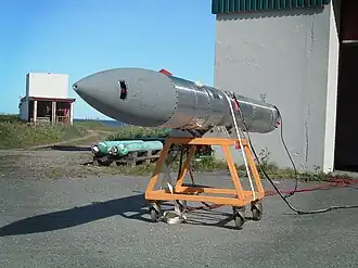 An elongated cyllindric object, ending with a cone, placed on a small, openwork trolley, in front of a hangar, on a sunny day.