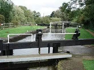 A canal lock. The lock chamber is full. Beyond the lock is a footbridge across the canal.