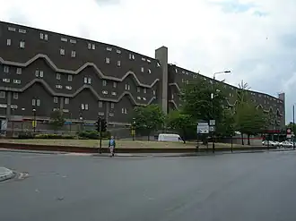 A long dark building with small windows and upper floors slightly overhanging lower ones. Two pale bands at the overhangs step up and down along the face of the building.