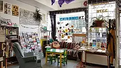 Sunlight shines through the window onto the hangout zone at Spartacus Books, which includes a couch, chair, guitar, colourful magazine rack, posters, and patches.