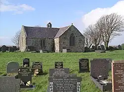 At the far end of a graveyard is a simple stone church, which appears L-shaped, and has a bellcote