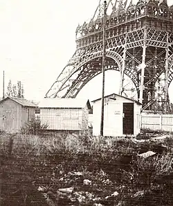 An old photograph of a series of small buildings resembling shacks. In the background is one of the legs of the Eiffel Tower