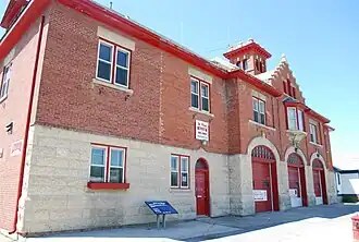 A two-storey concrete and red brick fire hall. There are three large doors for vehicles, one regular door, and two windows.