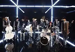 Ten Super Junior members wearing suit jacket in a black room with light above them.