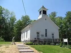 A small white church building, with trees in the background. A man walks towards the church.