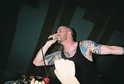 Vocalist Andrew Schwab performs at a concert. He has shaved his head and is wearing a black sleeveless shirt that displays his tattoos.