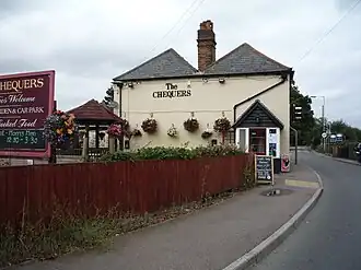 A photo of the Chequers Pub. It is a cream stucco two story wonky building on the bend in a road. A sign, saying "The Chequers" is displayed on the side of the building in a serif font.