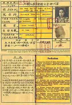 An old document made of one sheet of paper showing two outlined boxes, one in the top half and one on the bottom. The top box has writing in Japanese with a translation and a small head shot of a woman with a thumbprint below it. There are several small square imprints in red ink from a stamp. The bottom box is split into two vertical halves, one in Japanese and the other in Indonesian.