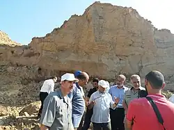 A group of ten people variously in dress shirts and polo shirts, and some wearing caps, stand in front of a yellow sandstone promontory.