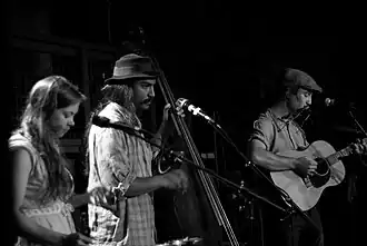A black-and-white image of three musicians (a woman and two men) playing instruments