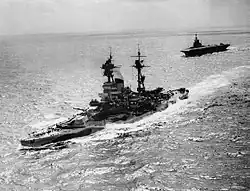 A large warship steaming through a calm sea with a large, flat-decked warship following behind