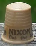 An off-white thimble, with the visible black lettering, "NIXON/Put the ne&nbsp;...