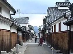 A narrow street lined by houses with a wooden lower part, a white upper storey and tile roofs.