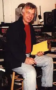 A gray-haired man with a red shirt and jacket sitting on a chair