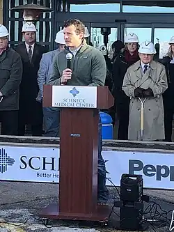 Hollingsworth, wearing a dark green jacket over a button-up shirt, speaks behind a podium with several people visible behind him in hard hats and long coats