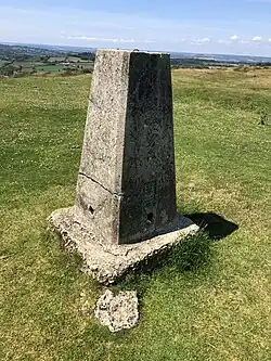 A triangulation stone at the summit of Pilsdon Pen. The stone is a neck height, thin stone column with markings of identification engraved in a copper emblem near the base. The stone sits in a field of grass and the Dorset landscape is in the background under a blue, slightly cloudy sky