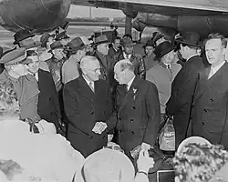 Truman and Attlee in dark suits shake hands. There are surrounded by a crowd of people, all dressed warmly. In the background is a propeller driven airliner.