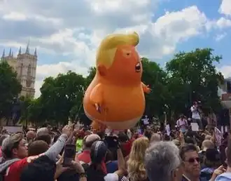 The helium balloon floating outdoors, held by restraining cords, surrounded by protesters, with the Houses of Parliament in the background