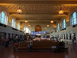 Interior of [[Union Station]] in [[New Haven|New Haven, Connecticut]], showing arched windows, hanging globe lights, digital departure boards, and passengers seated or walking through the concourse.