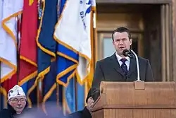 Senator Young, to the right side of the image, wears a suit while speaking in front a row of flags and at least one visible veteran