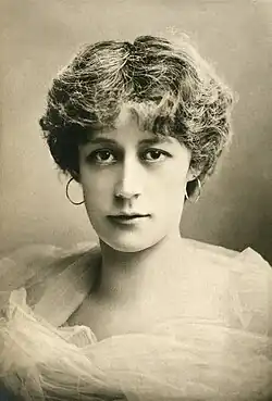 Head and shoulders of young white woman with dark hair, in a chiffon dress and hoop earrings, looking towards the camera
