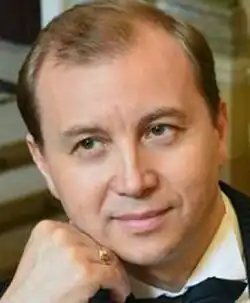 Colour publicity shot of a half-smiling, clean-shaven man with short, light brown hair, combed back. He is resting his chin on his hand.