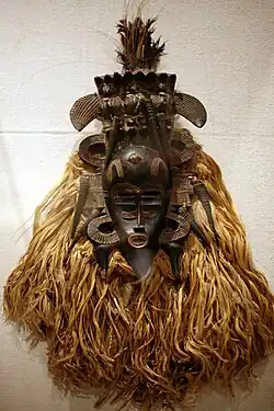 Poro mask; 19th-mid-20th century; wood, horns, raffia fiber, cotton cloth, feathers, metal; height: 301⁄4 in.; by Senufo people; Metropolitan Museum of Art. Designed to pay homage to female ancestors, this mask's serene dark oval face is offset by glinting brass, symmetrical extensions, and delicate patterns symbolizing wisdom and beauty