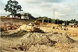 Photograph of construction of the Westgate Shopping Centre in Massey, Auckland, New Zealand, in 1994. The image depicts a pit of dirt with construction vehicles, including several trucks and a bulldozer.