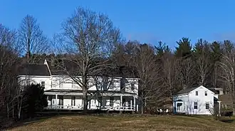 A long two-story white house with a black side-gabled roof and a tall bare tree in front of it seen from a distance. Next to it a much smaller side-gabled house, similar in architectural style, is seen from the side.