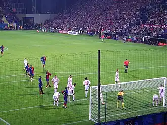 A photograph from behind the goal shows Barcelona and Wisła Kraków players gathering in the near penalty area for a corner kick. Wisła Kraków players are standing closer to the goal, and the referee is standing on the edge of the penalty area.