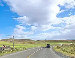 A road through an open landscape of green grass and brownish green hills in the distance