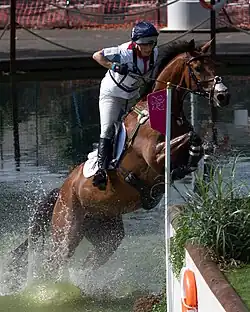 white-attired rider on a chestnut horse which is leaping out of a water jump