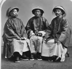 A black and white photo of three men in Chinese official uniform seated. They all wear hats with tassels.