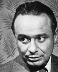 Black and white, dramatic-pose publicity shot, of a clean-shaven man with designer-stubble moustache, big dark eyes, and receding, oiled black hair.