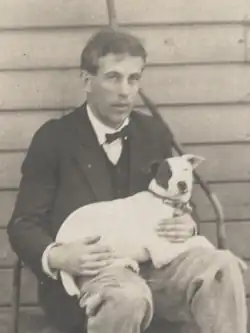 James Arbuthnot, sitting on a sod roller. He's dressed in rough trousers, and coat over a white shirt and vest. His dog is laying across his lap, hind legs on Arbuthnot's right leg. The dog is white save for a dark patch around its right eye.