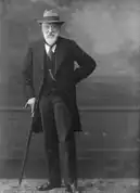 An elderly bearded man in a light hat wearing a suit propping himself up with a cain