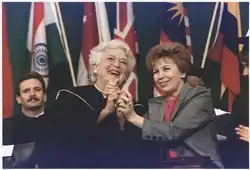 Barbara Bush and Raisa Gorbacheva stand smiling and clasping each other's hands. Bush is wearing a graduation robe.